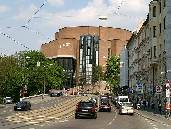 A public road during the day with car on the road and people walking on footpath. in the background is the Gasteig, the philharmony of Munich.