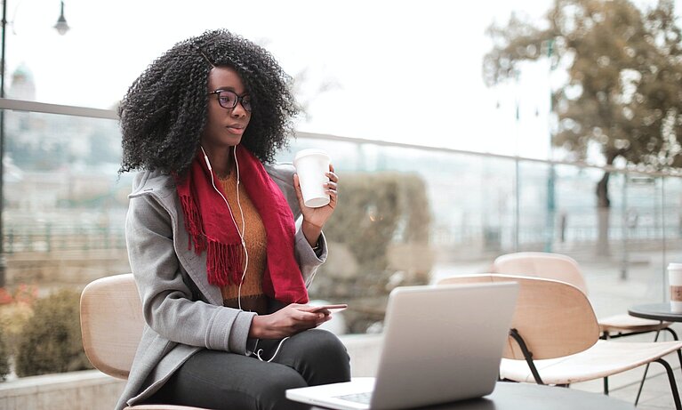 Afro american student is sitting on a terrace, drinking coffe and working on laptop